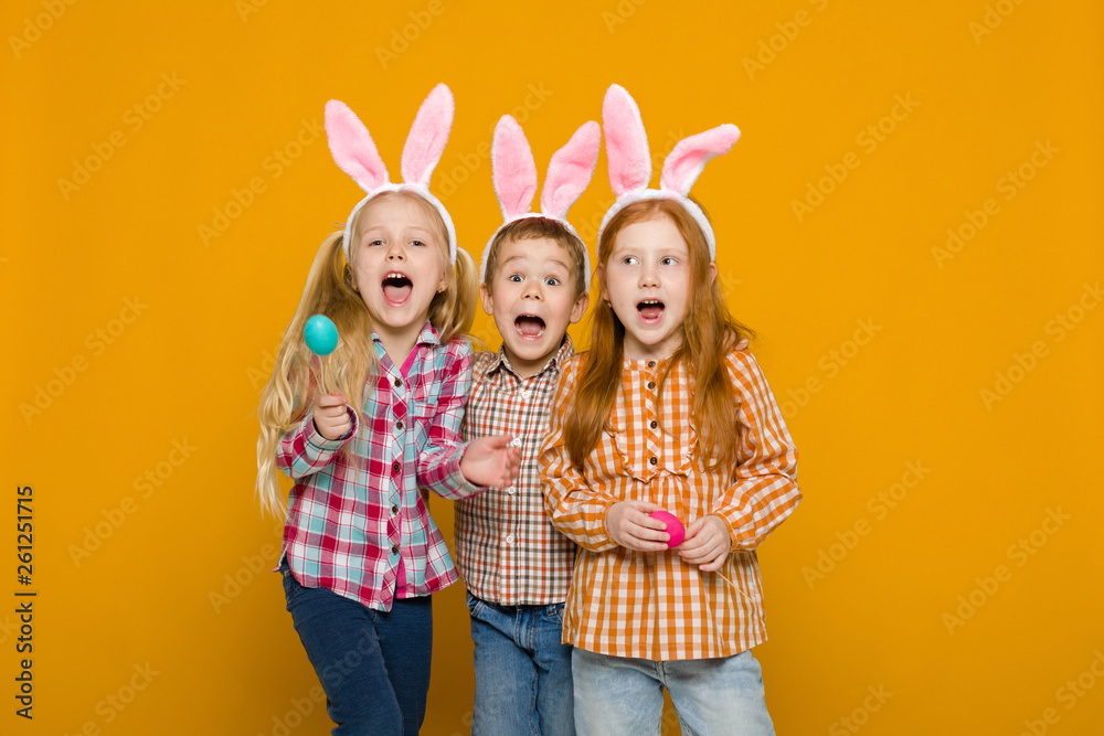 surprised two little girls and boy with Easter bunny ears holding colorful eggs on yellow background. Happy easter. cute children surprised two little girls and boy with Easter bunny ears holding colorful eggs on yellow background. Happy easter. cute children