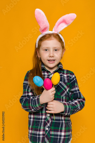 Portrait of cute red-haired little child girl with Easter bunny ears holding colorful eggs on yellow background. Happy easter