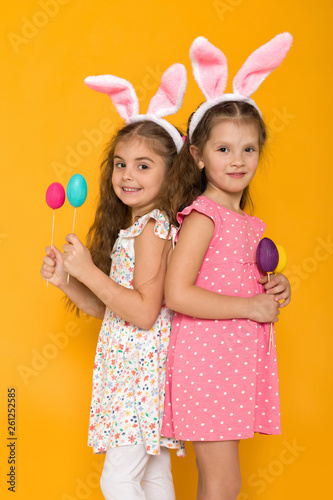 two little girls with Easter bunny ears holding colorful eggs on yellow background. Happy easter. cute children