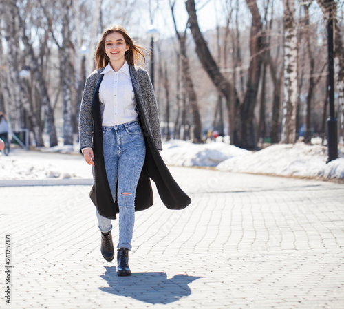Brunette girl in a gray coat and blue jeans walks in spring park © Andrey_Arkusha