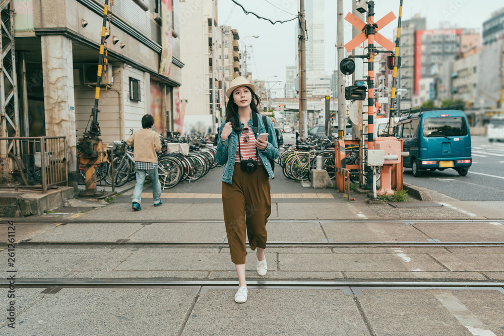 Fototapeta premium full length trendy young asian woman traveler walking outdoors holding mobile phone texting online message. beautiful female backpacker carrying professional camera while crossing railway osaka japan