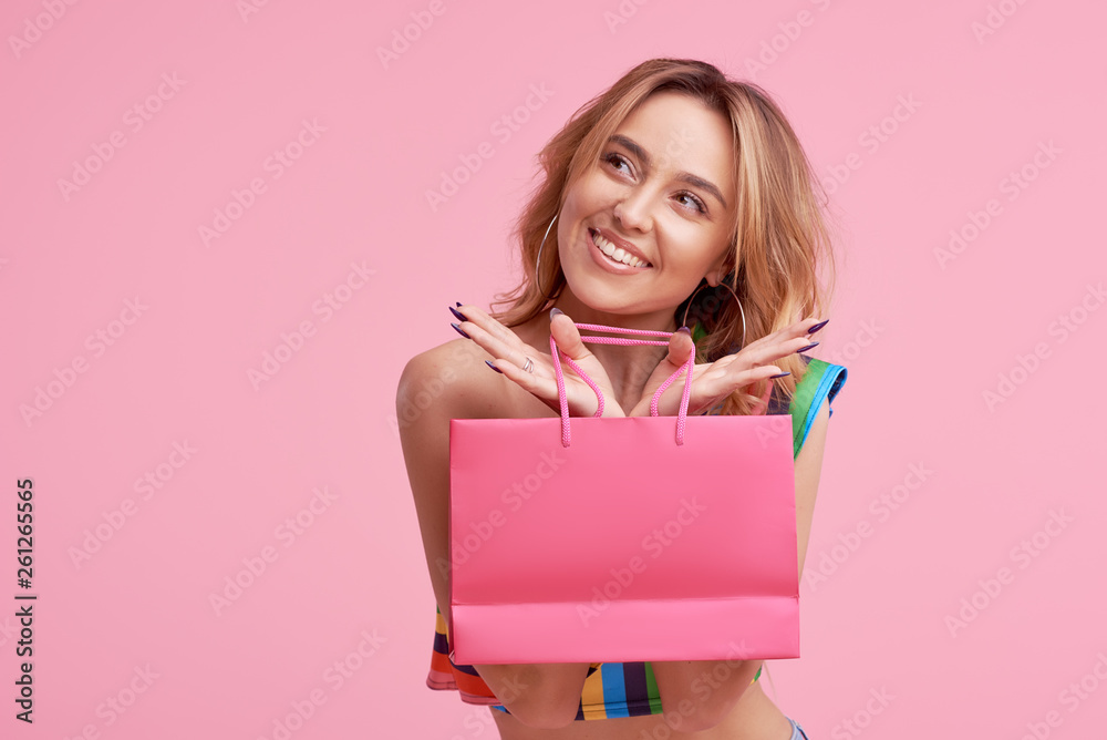 Satisfied buyer. Portrait of an excited happy girl wearing colorful summer clothes holding shopping bag isolated over pink background. Place to sign or text on the package. Sale concept Satisfied buyer. Portrait of an excited happy girl wearing colorful summer clothes holding shopping bag isolated over pink background. Place to sign or text on the package. Sale concept