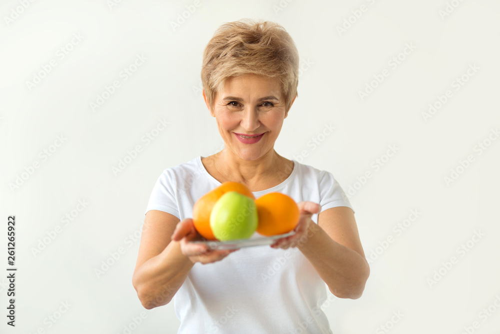 beautiful aged woman with a short haircut in a white T-shirt with a plate of fresh fruit on a white background beautiful aged woman with a short haircut in a white T-shirt with a plate of fresh fruit on a white background