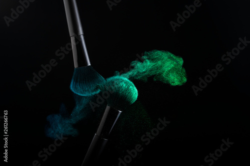Cosmetic shades of different colors, blue and green, fly away from two make-up brushes creating a fancy pattern on a black background.