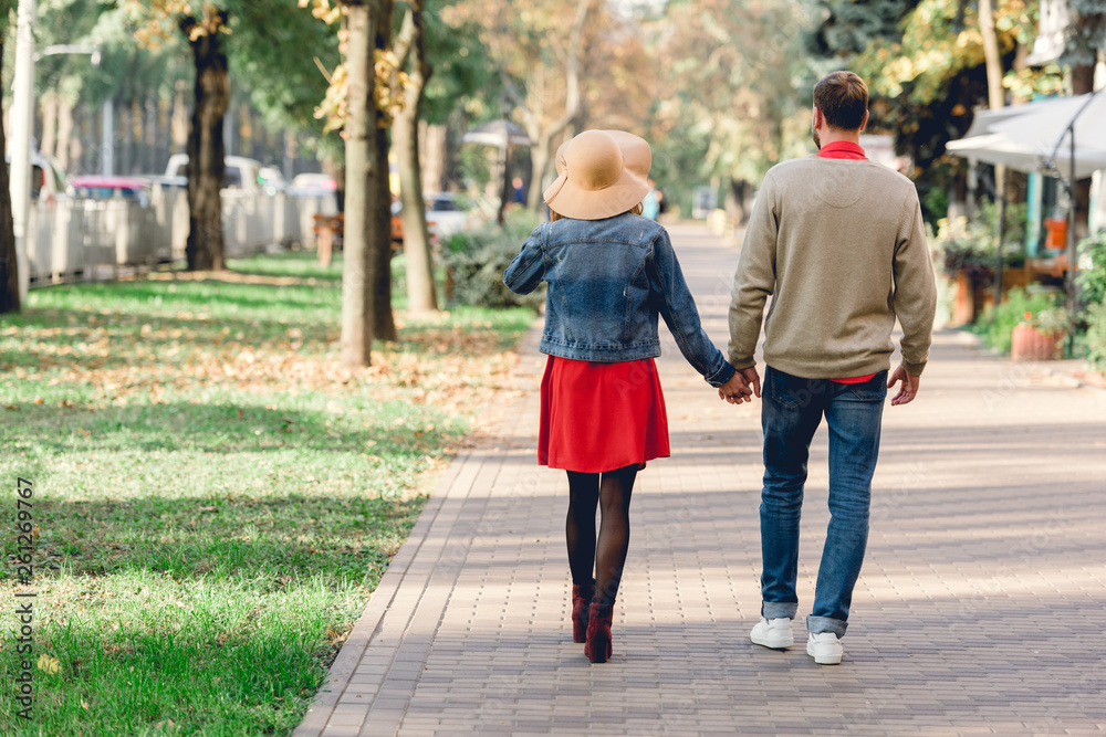 back view of man and woman in hat holding hands while walking in park
