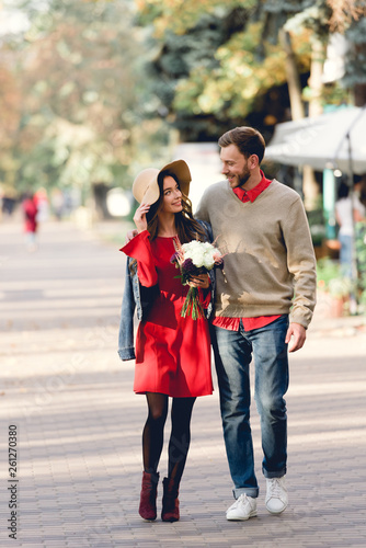 happy man looking at girlfriend in hat holding flowers while walking in park