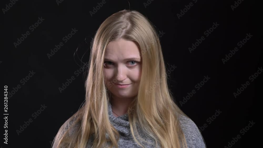 Portrait of young blonde straight-haired model watching shyly into camera on black background.