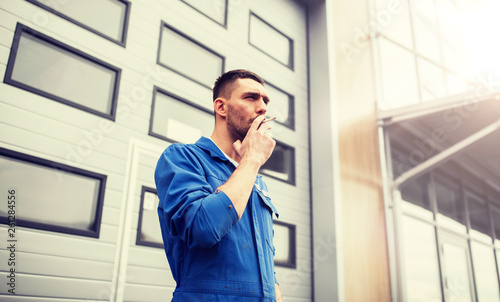 service, repair, maintenance and people concept - auto mechanic smoking cigarette at car shop or garage outdoors