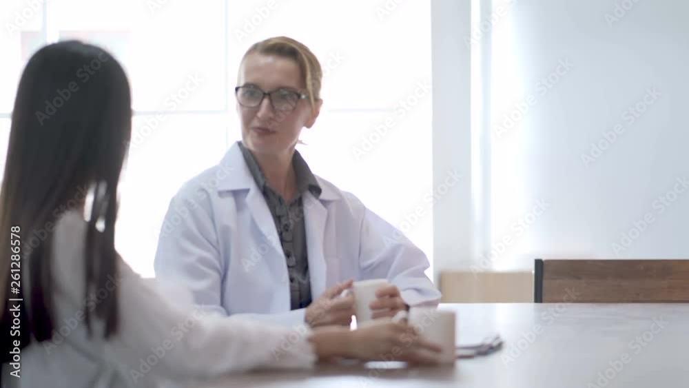 The senior female doctor wearing lab coat drinking hot coffee, smiling and talking with colleagues while having a break in clinic