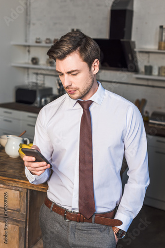 Φωτογραφία handsome confident man holding smartphone while standing in kitchen at home