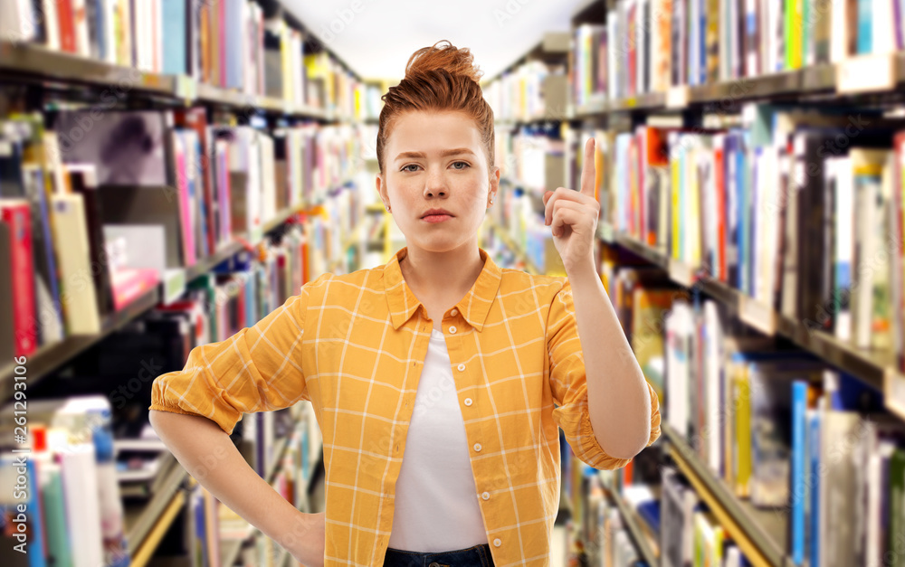 gesture, counting and people concept - serious red haired teenage student girl in checkered shirt pointing finger up over library background gesture, counting and people concept - serious red haired teenage student girl in checkered shirt pointing finger up over library background