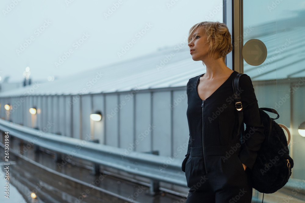 A woman with a city backpack is thoughtfully standing near glass doors. A woman with a city backpack is thoughtfully standing near glass doors.