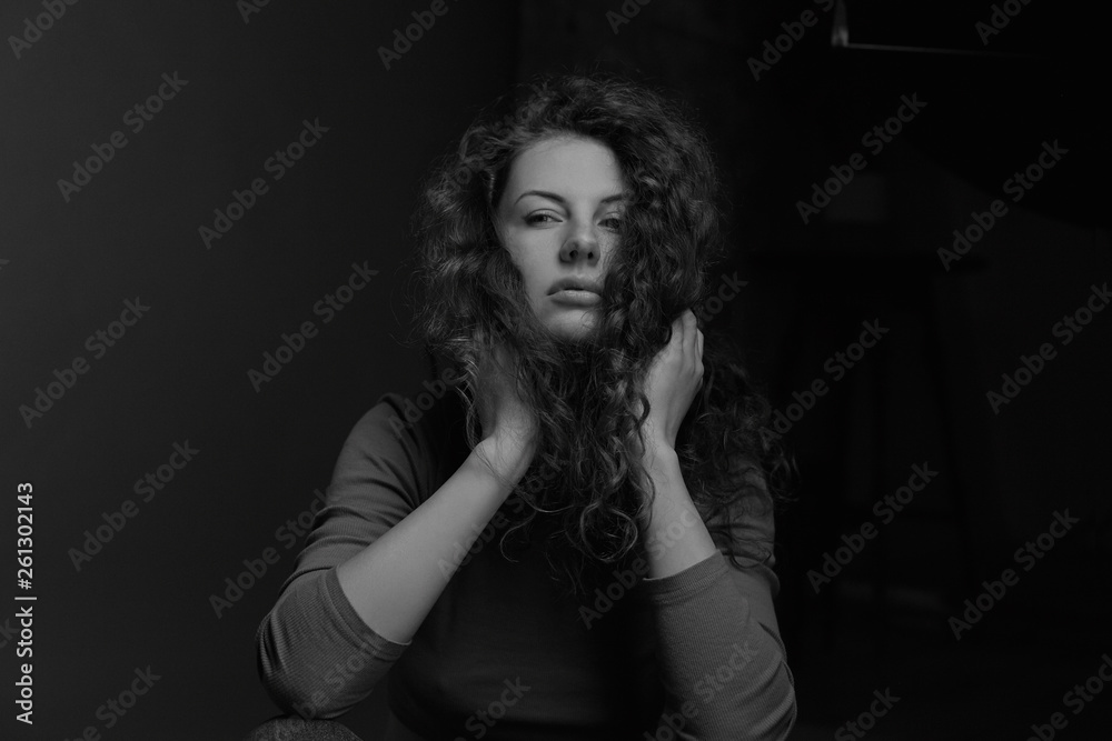 Plakat Black and white authentic horizontal portrait of thoughtful Caucasian curly haired female model, holding hands on chin, mysteriously looking sideways, posing in dark room indoors. Beauty concept.