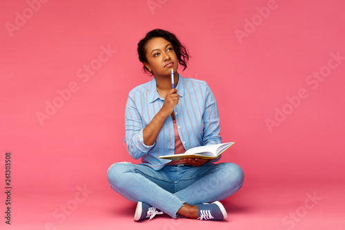education, high school and inspiration concept - african american young student woman with pen and diary or notebook sitting on floor and thinking over pink background