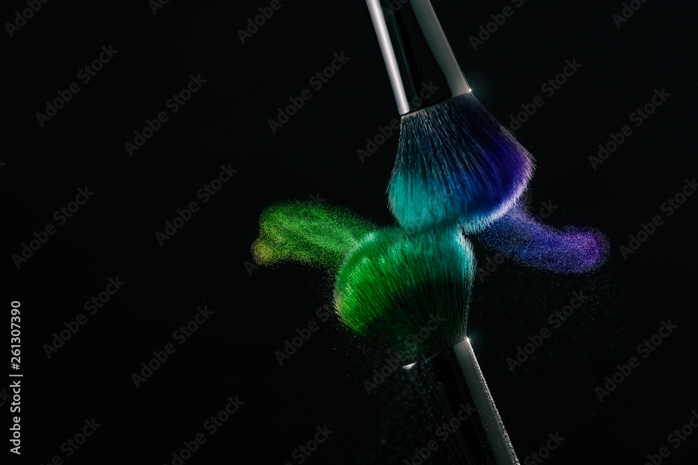 Cosmetic shades of different colors, blue and green, fly away from two make-up brushes creating a fancy pattern on a black background.