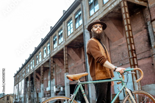 Lifestyle portrait of a bearded hipster dressed stylishly walking with retro bicycle on the industrial urban background
