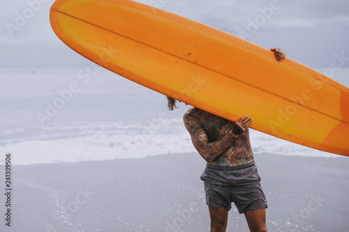 Canvas Print Attractive male surfer in tattoos on the ocean with an orange bright longboard surfboard
