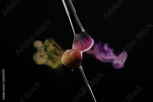 Cosmetic shades of different colors, yellow and pink, fly away from two makeup brushes creating a fancy pattern on a black background.