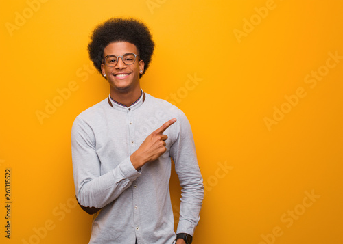 Young african american man over an orange wall smiling and pointing to the side