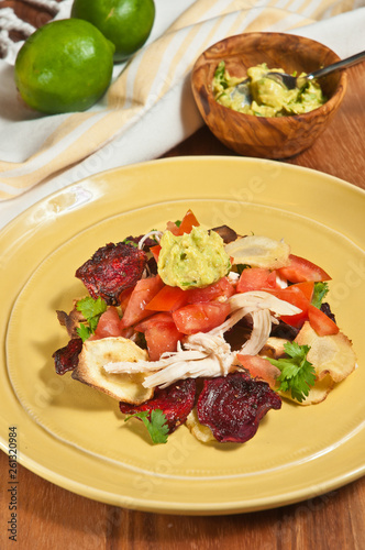 Top view, medium distance of sliced beets, parsnip chip nachos with shredded chicken, sliced tomato, guacamole, parsley on a round, yellow plate- paleo diet