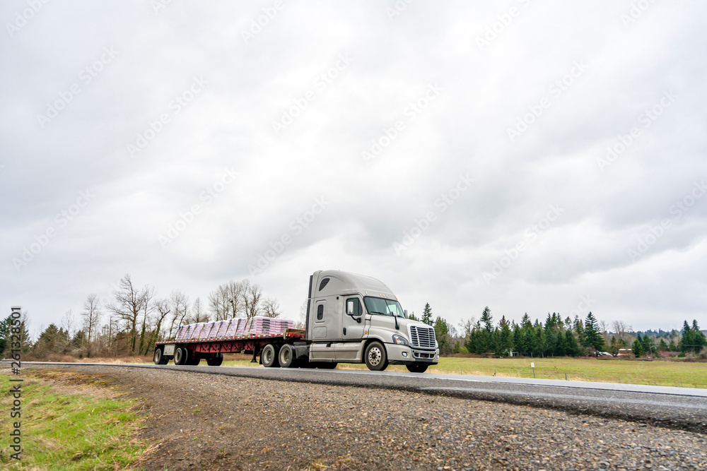 Big rig semi truck transporting fastened commercial cargo on flat bed semi trailer going on the road in overcast weather