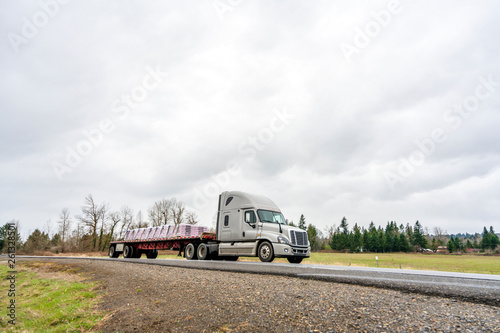 Big rig semi truck transporting fastened commercial cargo on flat bed semi trailer going on the road in overcast weather