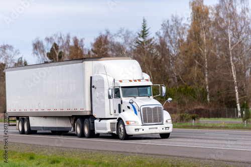 Long haul white big rig semi truck transporting goods in dry van semi trailer running on straight highway road