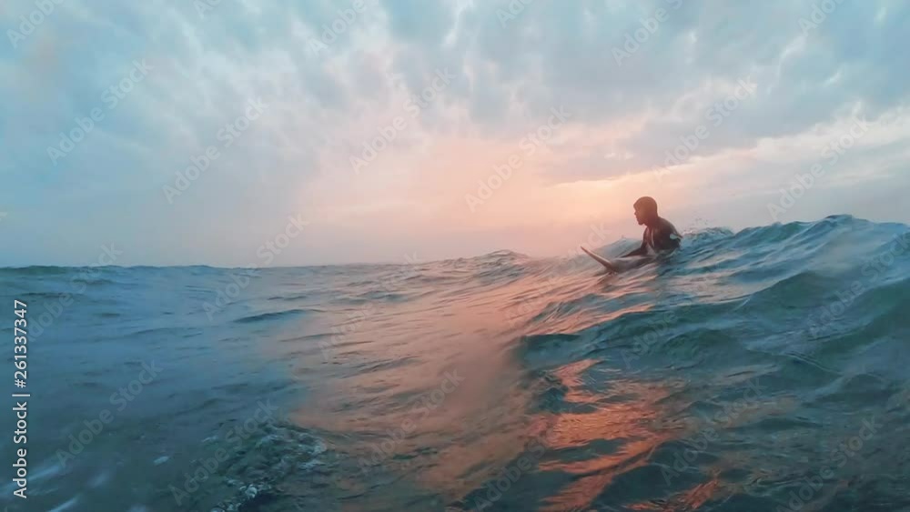 A cold water surfer is waiting in the line up in the ocean in a beautiful sunset while a wave approaches. He does a duck dive. 