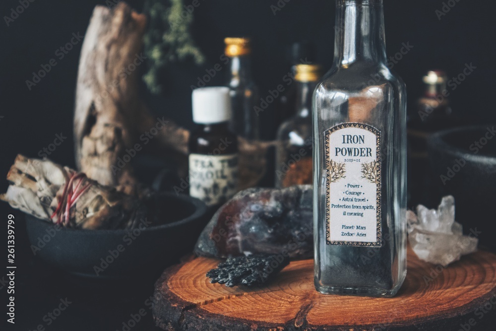 Close up of a bottle of iron dust powder with vintage apothecary label on witch altar. Various spell ingredients in background - glass jars, bottles, sage smudge stick in cast iron holder, crystals