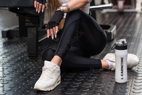Attractive model sits with L-carnitine drink in modern gym. Lower part view photo