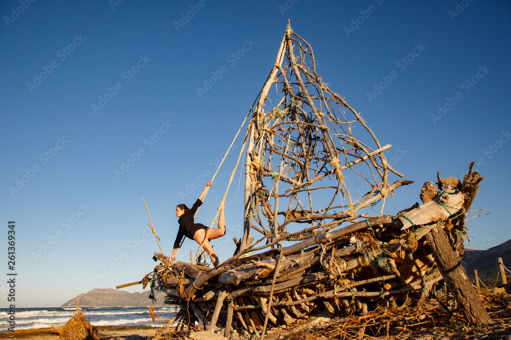 Back view of young dancer with upped leg posing on wooden boat on sand coast with waving sea and blue sky Back view of young dancer with upped leg posing on wooden boat on sand coast with waving sea and blue sky