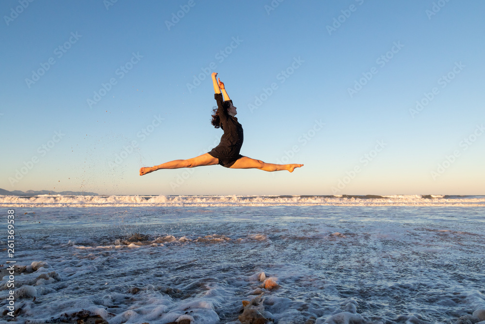 Side view of young dancer in air with legs to sides on sand coast with waving sea and blue sky Side view of young dancer in air with legs to sides on sand coast with waving sea and blue sky