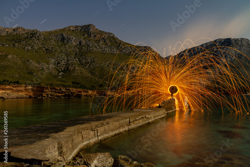 Long exposure of silhouette of human with burning round and fireworks on rocks between water near mountains in evening