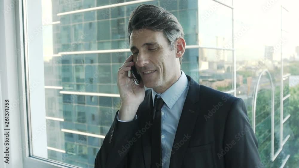 Zoom in shot of smiling mature businessman talking on smartphone while standing near office window Zoom in shot of smiling mature businessman talking on smartphone while standing near office window