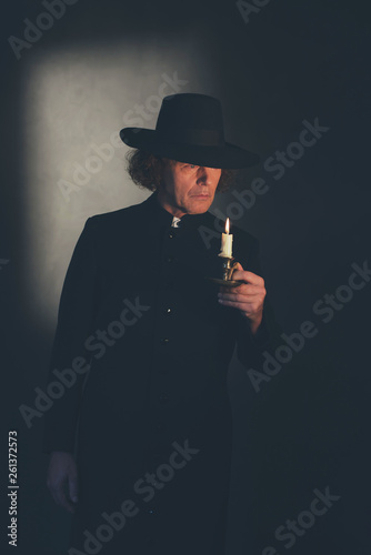 Mysterious victorian priest in black coat and hat holding candlestick.