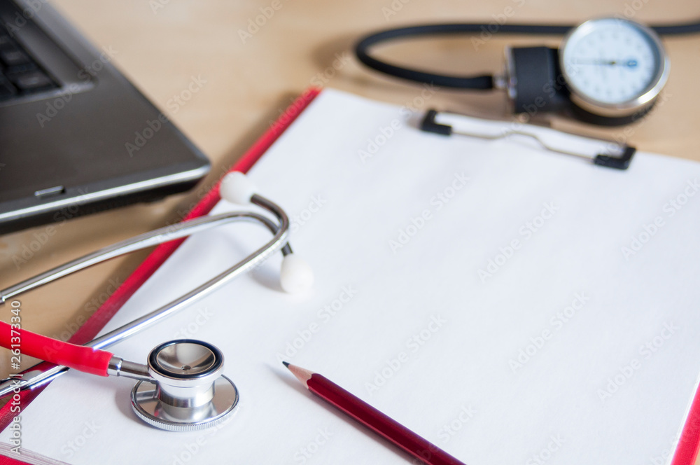 Red stethoscope and red pencil on a red clipboard on a table. Near laptop and tonometer. Copy space. Medical device. Treatment, health care. Heart examination. Studying the pulse. Close-up. Red stethoscope and red pencil on a red clipboard on a table. Near laptop and tonometer. Copy space. Medical device. Treatment, health care. Heart examination. Studying the pulse. Close-up.