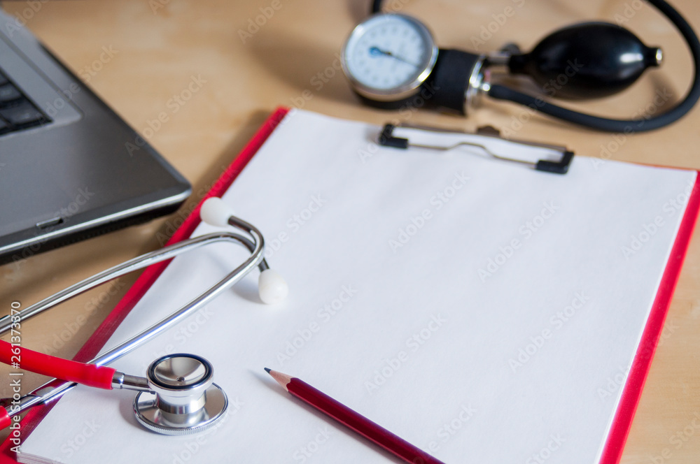 Red stethoscope and red pencil on a red clipboard. Near laptop and tonometer. Copy space. Medical device. Treatment, health care. Heart examination. Studying the pulse. Red stethoscope and red pencil on a red clipboard. Near laptop and tonometer. Copy space. Medical device. Treatment, health care. Heart examination. Studying the pulse.