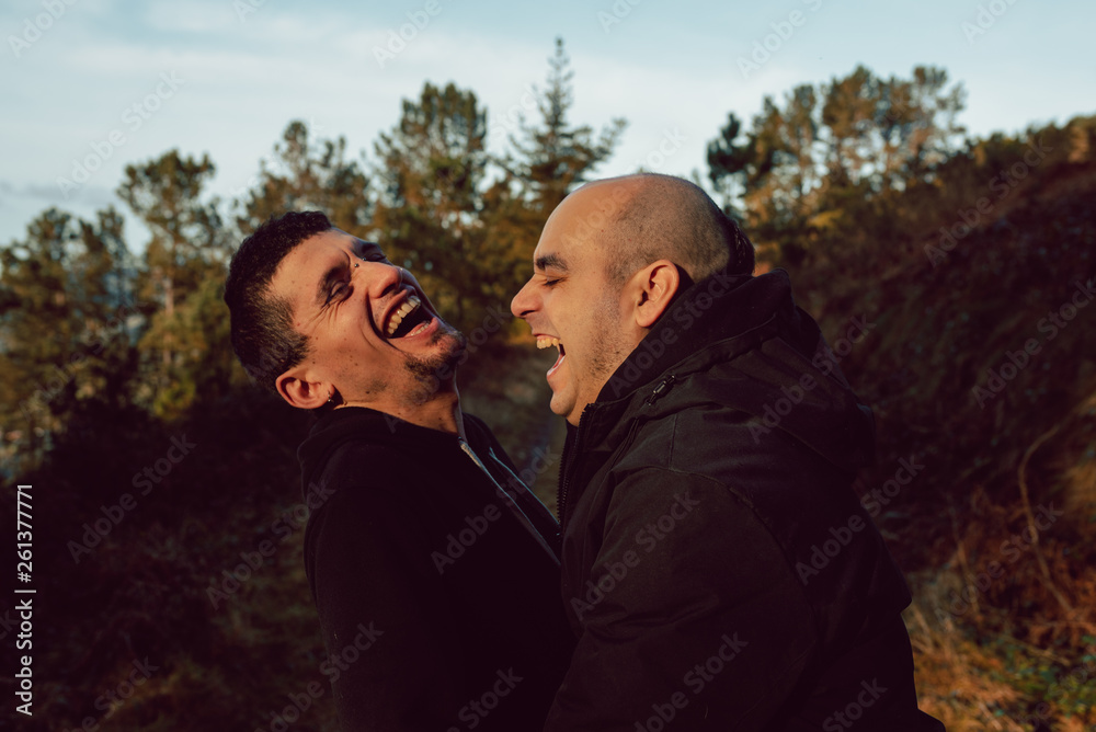 Side view of cheerful homosexual couple embracing and looking at each other on route in forest in sunny day Side view of cheerful homosexual couple embracing and looking at each other on route in forest in sunny day