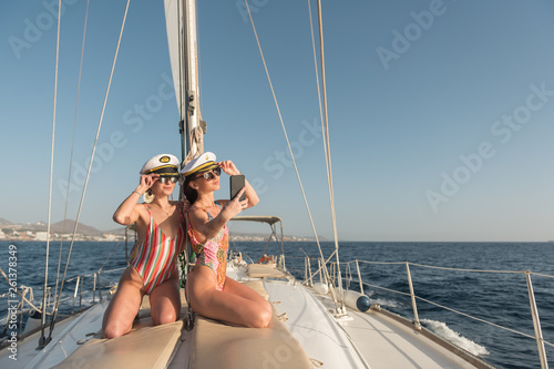 Beautiful young females in sunglasses and captain hats taking selfie on mobile phone and sitting on deck of expensive boat floating on water in sunny day photo