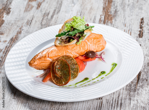 Baked salmon garnished with olives, greens, tomatoes on plate over wooden background. Hot fish dish