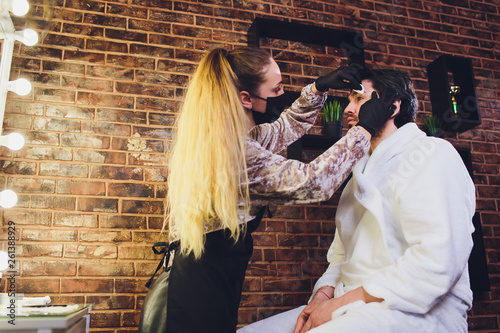 Professional barber in white shirt doing threading procedure and correcting shape of eyebrows to young male client sitting in chair in barber shop. Concept of styling and care. photo