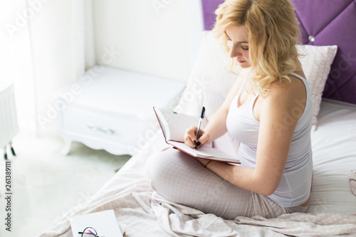 Attractive blonde woman sits on the edge of the bed at home and writes into a notebook