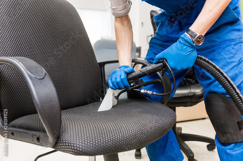 Young man in workwear and rubber gloves cleans the office chair with professional equipment.