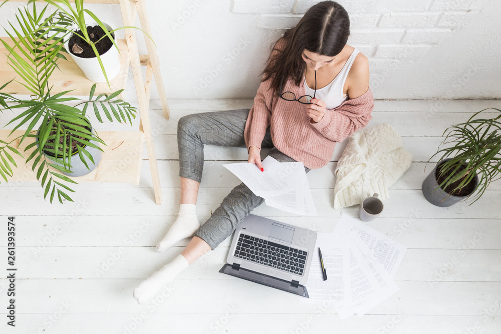 A young happy woman sits on the floor in a bright apartment or office interior and works at a laptop, freelancer girl at work, the concept of distance learning students, paperwork and online learning A young happy woman sits on the floor in a bright apartment or office interior and works at a laptop, freelancer girl at work, the concept of distance learning students, paperwork and online learning