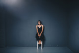 social theme of female loneliness is pain suffering. Abastration of male domestic violence. Young beautiful Caucasian woman in black clothes, strange pose, white chair big dark room. Fear and despair