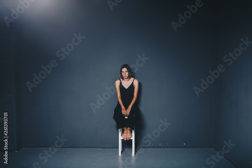 social theme of female loneliness is pain suffering. Abastration of male domestic violence. Young beautiful Caucasian woman in black clothes, strange pose, white chair big dark room. Fear and despair