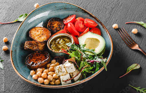Healthy Buddha bowl dish with avocado, tomato, cheese, chickpea, fresh arugula salad, baked potatoes and sauce pesto in black background. Dieting food, clean eating. © Jukov studio