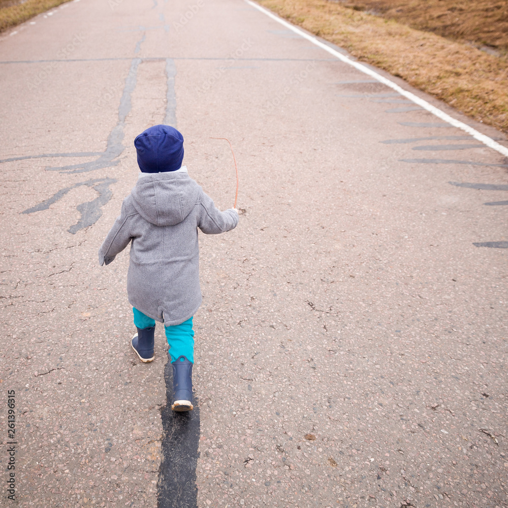 A little boy walks on an asphalt road in rubber boots, jeans and a ...