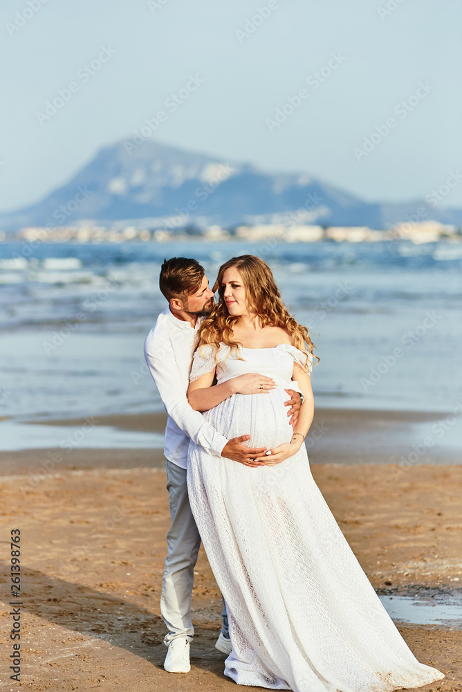 Young man hugging his pregnant wife in white dress on a beach Young man hugging his pregnant wife in white dress on a beach