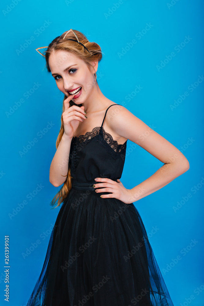 Portrait of beautiful woman on blue background, young lady posing at party, masquerade, carnival, biting finger. Wearing fancy black dress, cat ear headband in diamonds, gold manicure. Portrait of beautiful woman on blue background, young lady posing at party, masquerade, carnival, biting finger. Wearing fancy black dress, cat ear headband in diamonds, gold manicure.